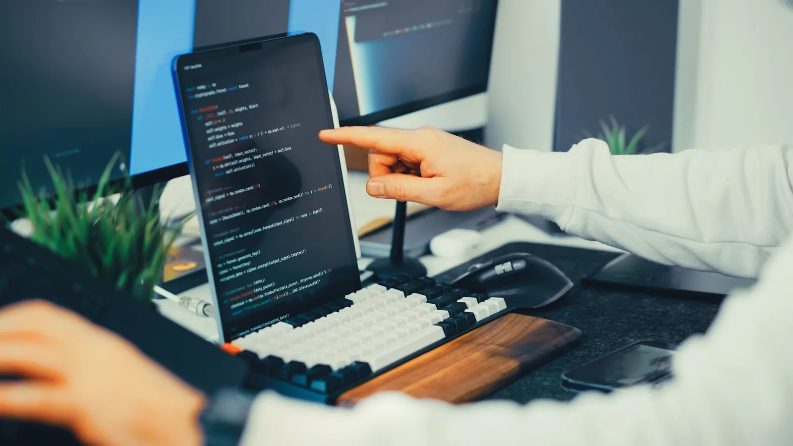 A developer pointing at code on a tablet propped above a keyboard, with two monitors behind it